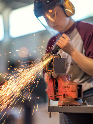 Sparks fly as student wearing protective helmet and earmuffs touches metal to grinding machine. 