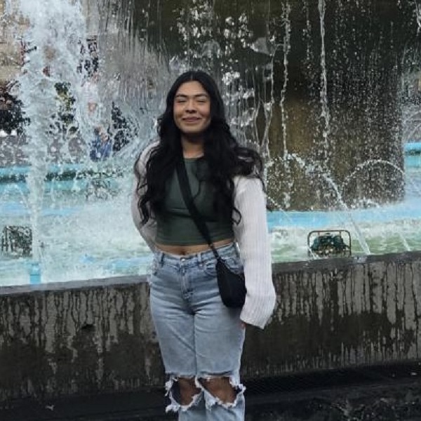 Young lady with long brown wavy hair wearing a green top, blue jeans, and a white sweater standing in front of a water fountain.