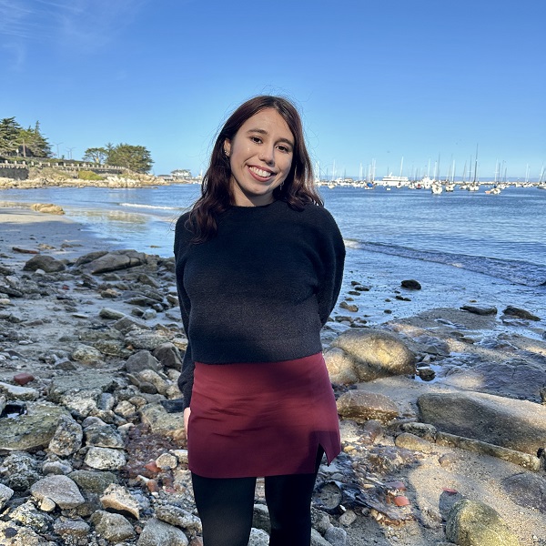 Young lady with long brown wavy hair and brown eyes wearing a black sweater, burgundy skirt, and black leggings standing on a rock beach.