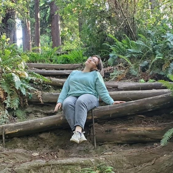 Emilyann wearing a teal sweatshirt and jeans, with shoulder length light brown hair, sitting on a log in the woods