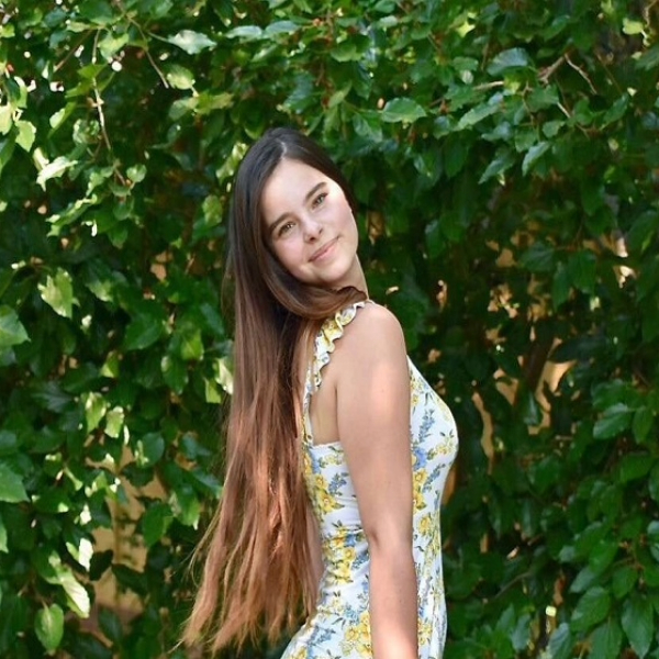 Meadow with long straight brown hair, wearing a white sundress with yellow and blue flowers, standing in front of a tall green leafy bush