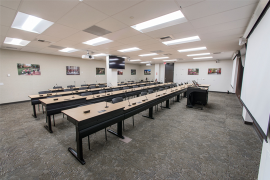 Rows of desks with microphones and powercords face large projector screens