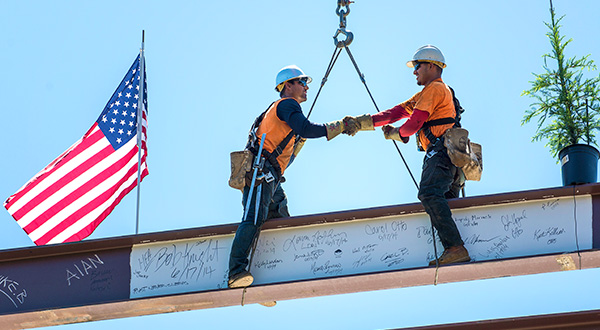 Construction workings shake hands high up on beams with American flag waving in the background