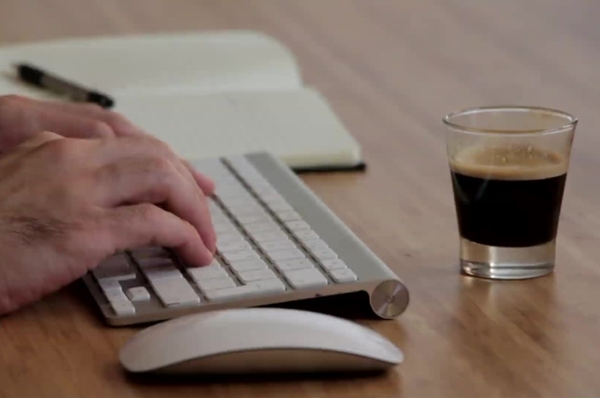Hands typing at a keyboard with a shot of espresso next to the keyboard.