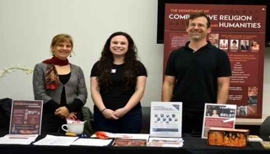 three faculty staff members standing at a table, smiling, at a tabling event