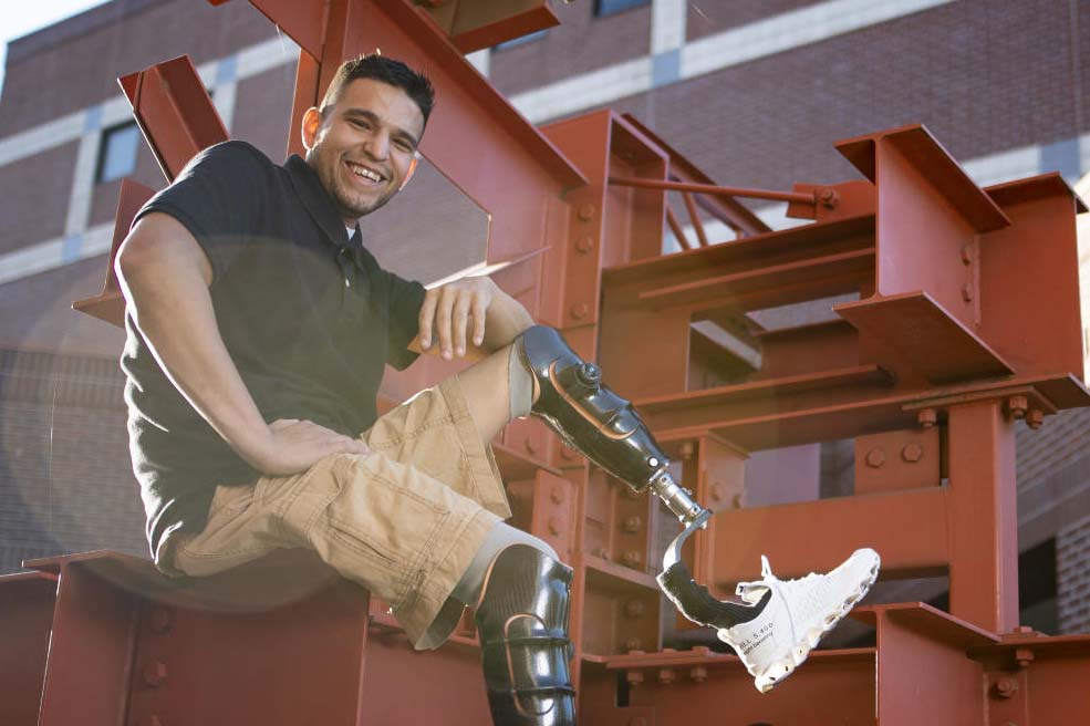 A student with prosthetic legs poses with them kicked up on a campus sculpture