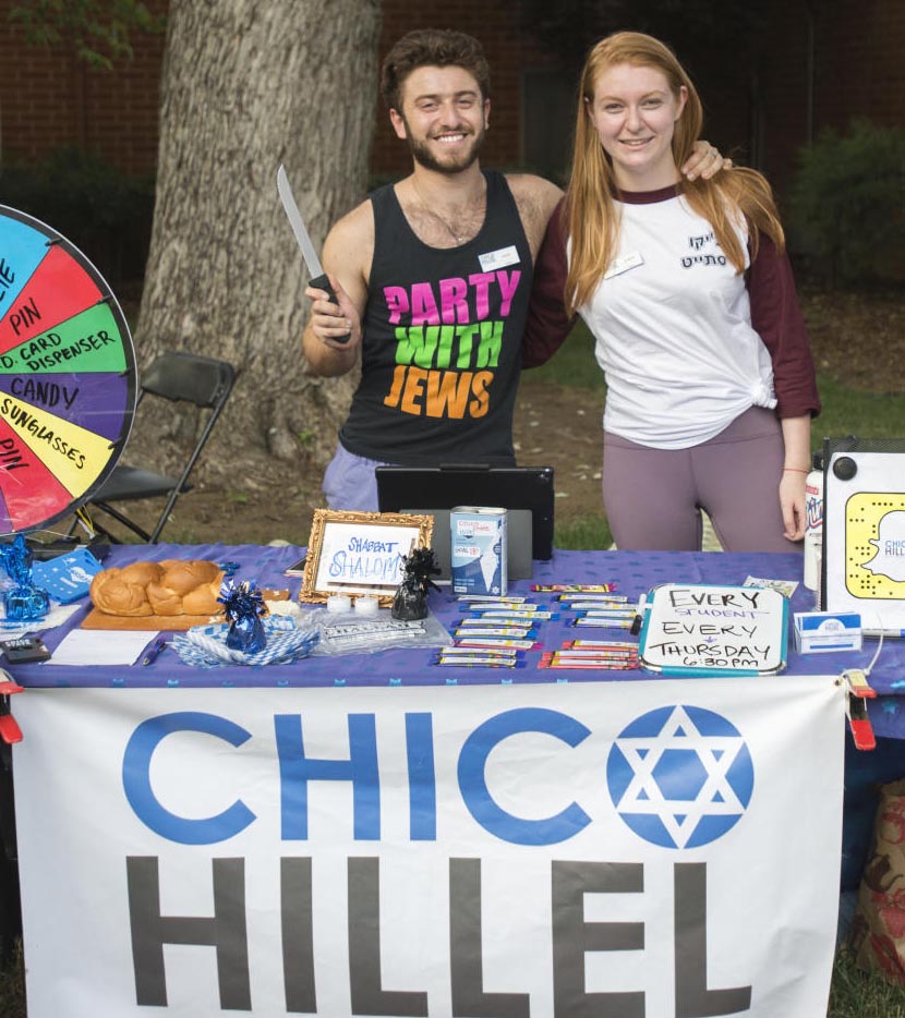 Students pose in front of a Chico Hillel sign at an event. One student is wearing a tank top that reads, Party with Jews