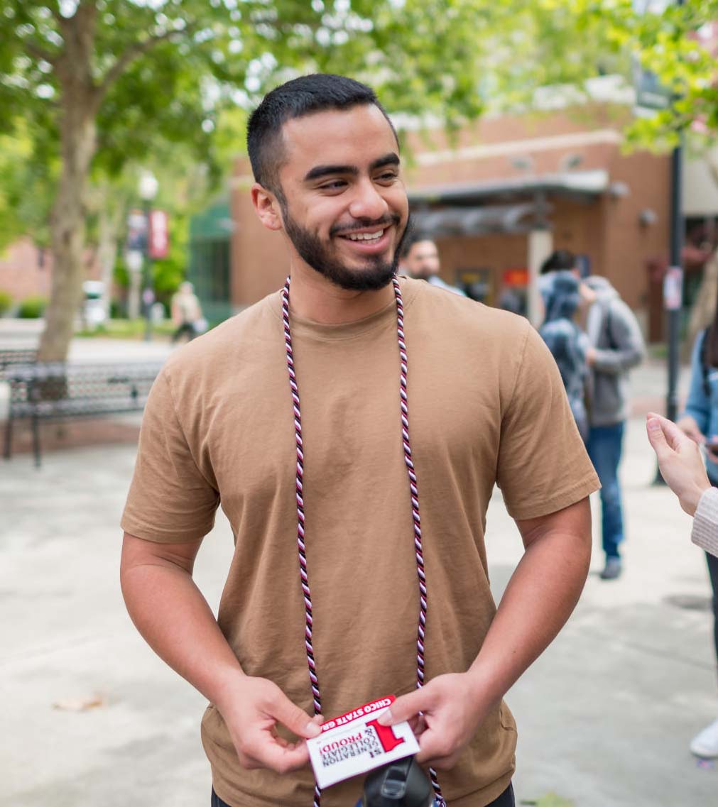 A student wears a grad cord for first gen students and holds a "1st Gen and Proud" sticker