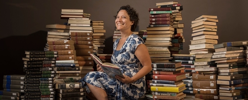 Professor Maitreya Badami sitting on books, surrounded by books, holding a book.