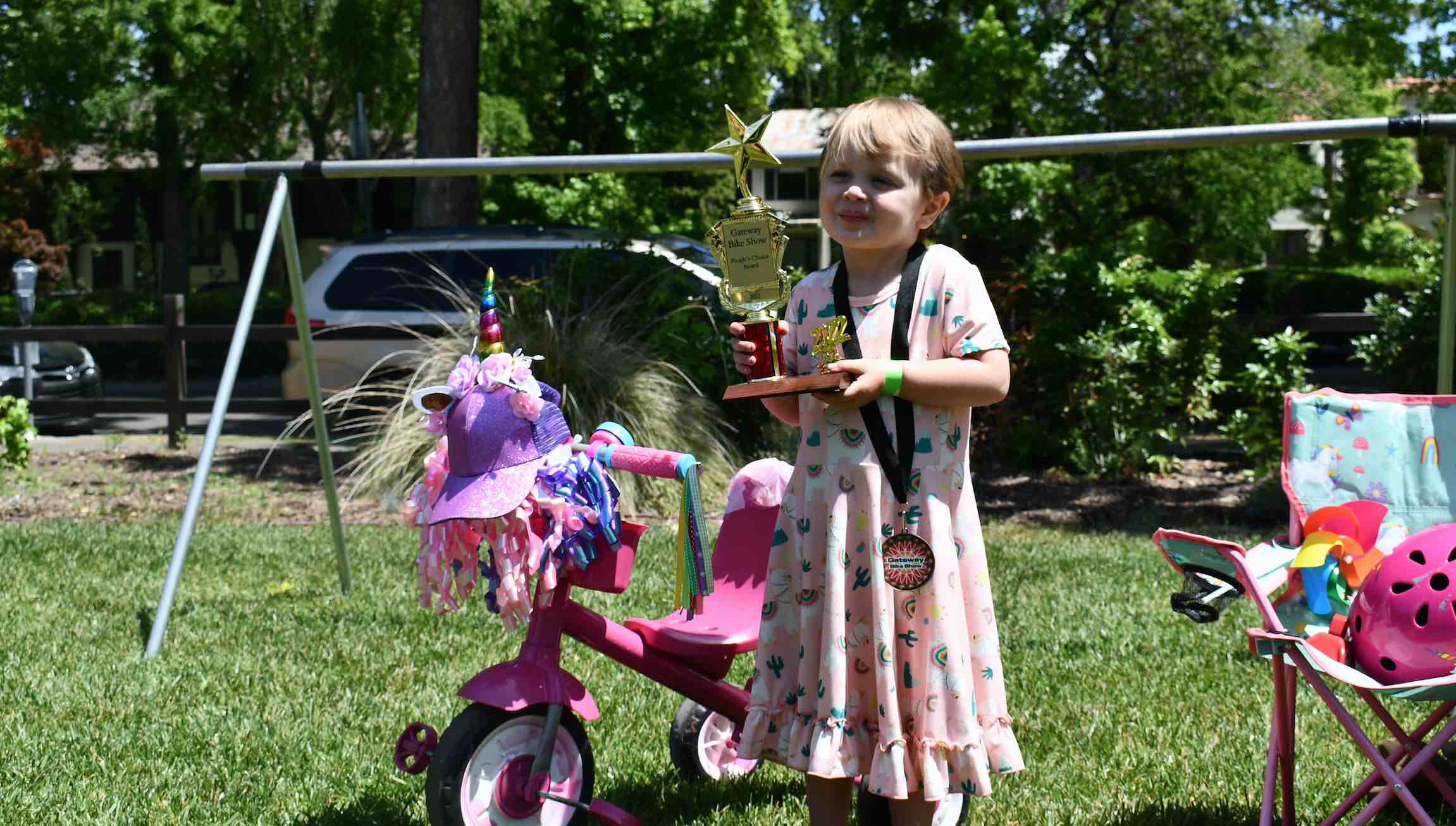 A young child with short blond hair and a pink dress stands next to their pink tricycle. They are holding a trophy