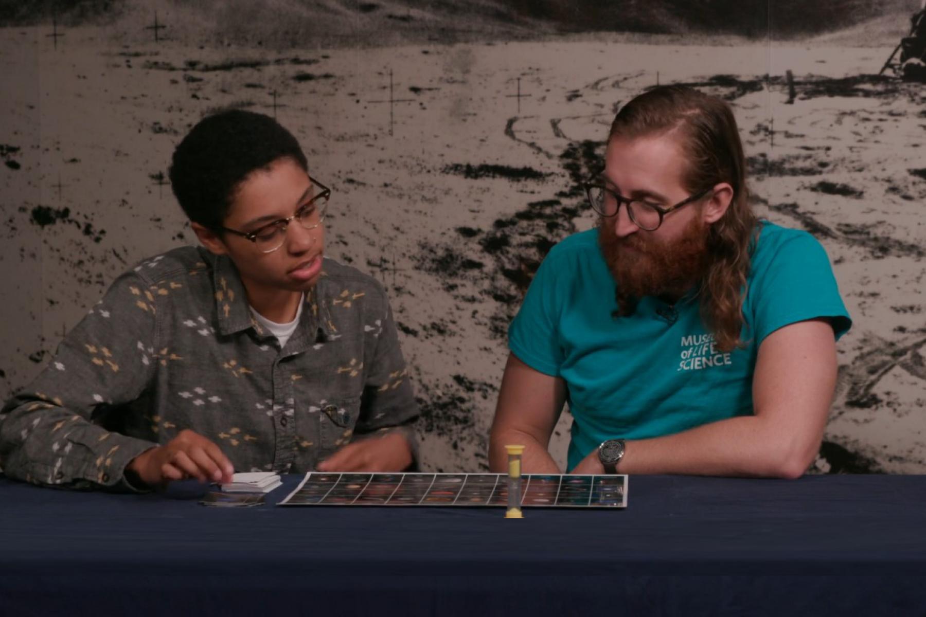 An adult and young adult sit at a table and play a science game