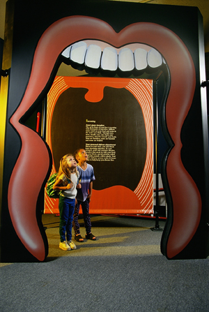 Two kids stand under an open mouth at the Microbes exhibit