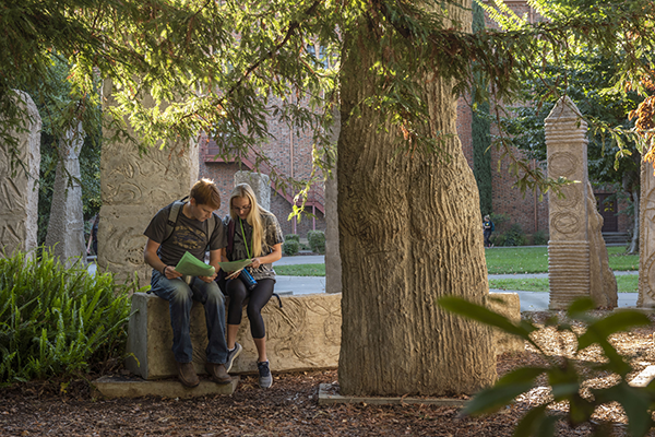 Two students review homework beside a series of sculptures that look like ancient standing stones, The Monoliths (Deborah Masters - 1999) are located beside Ayres Hall 