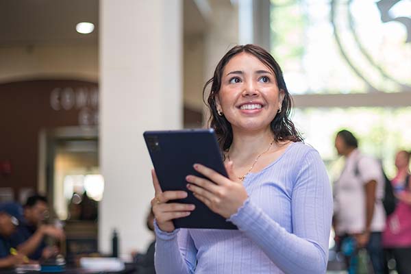 Holding a new tablet supplied by a Technology Equity and Inclusion event, a smiling student stands on campus