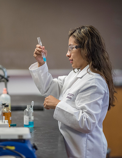 A female student in a lab coat gazes into a glass beaker in a science lab. 