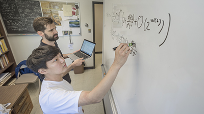 A student works out an equation on a whiteboard as instructor supervises
