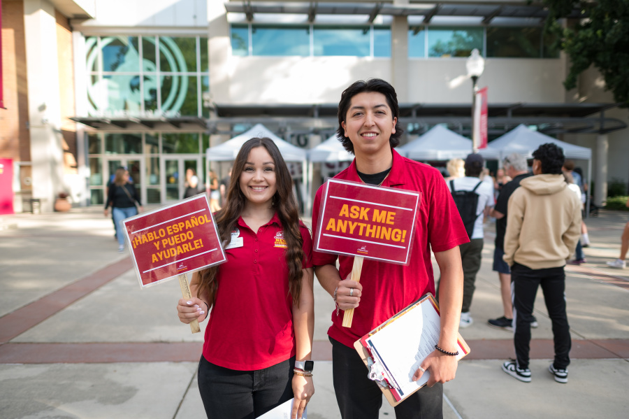 Two student orientation workers hold up signs in English and Spanish that encourages the viewer to ask for help.