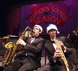 2 male students playing instruments and wearing santa hats in front of "Glorious Sounds of the Season" backdrop