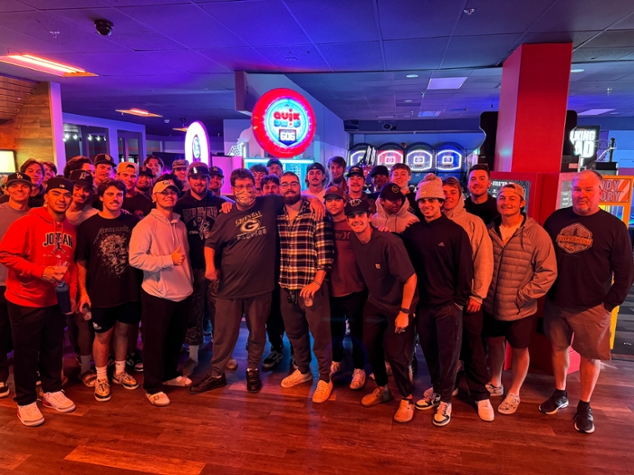 The whole baseball team at a bowling alley with their arms around a man wearing a mask. man