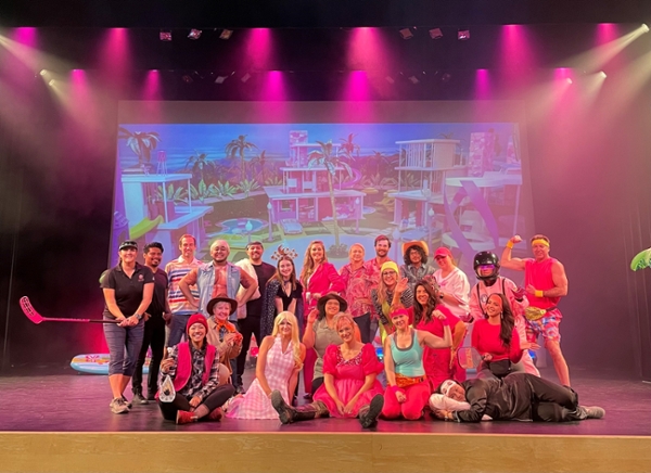 24 staff of Associated Students on the BMU Stage dressed as various versions of Barbie and Ken, including workout Barbie, sporty Barbie, weird Barbie, slumber party Barbie, and the Kens include a couple of real and fake muscle shirts, preppy clothes, and a cowboy hat among others. The backdrop behind them is a scene of several Barbie Dream houses in front of the ocean, presumably a shot from the Barbie movie.