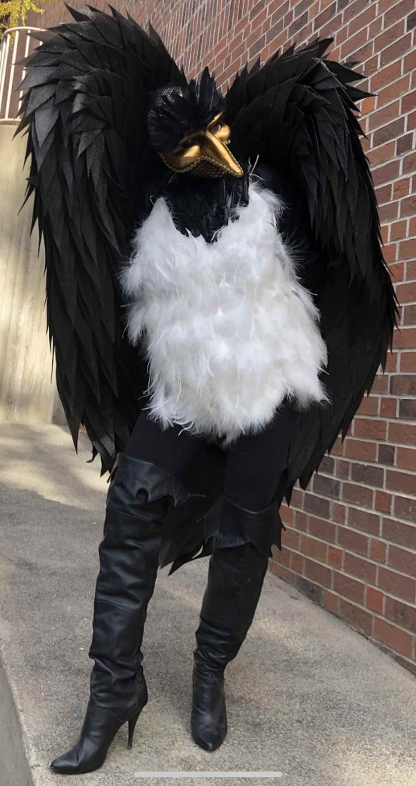 Renee Boyd dressed as a yellow billed magpie.  She is wearing a venetian gold mask with a beak.  Her front is covered with white feathers; she has two beautifully constructed wings arching overhead covered in black feathers, and she's wearing thigh high black leather boots.