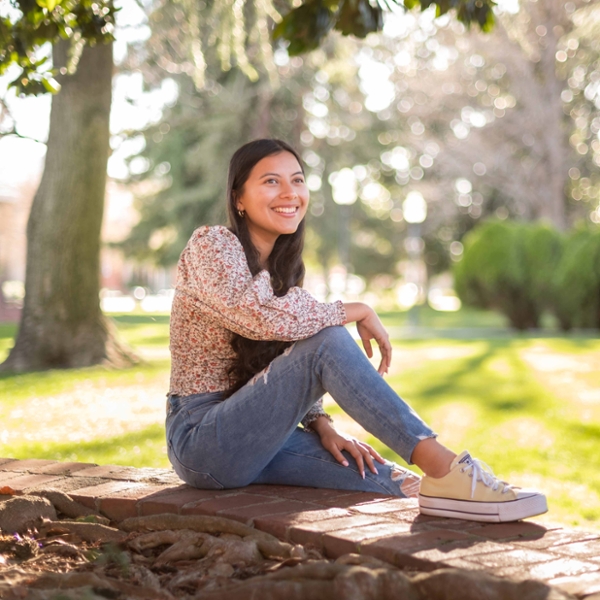 Student Yarely Contreras sits with one foot up on a tree retaining wall made of bricks as she looks onto into the distance.